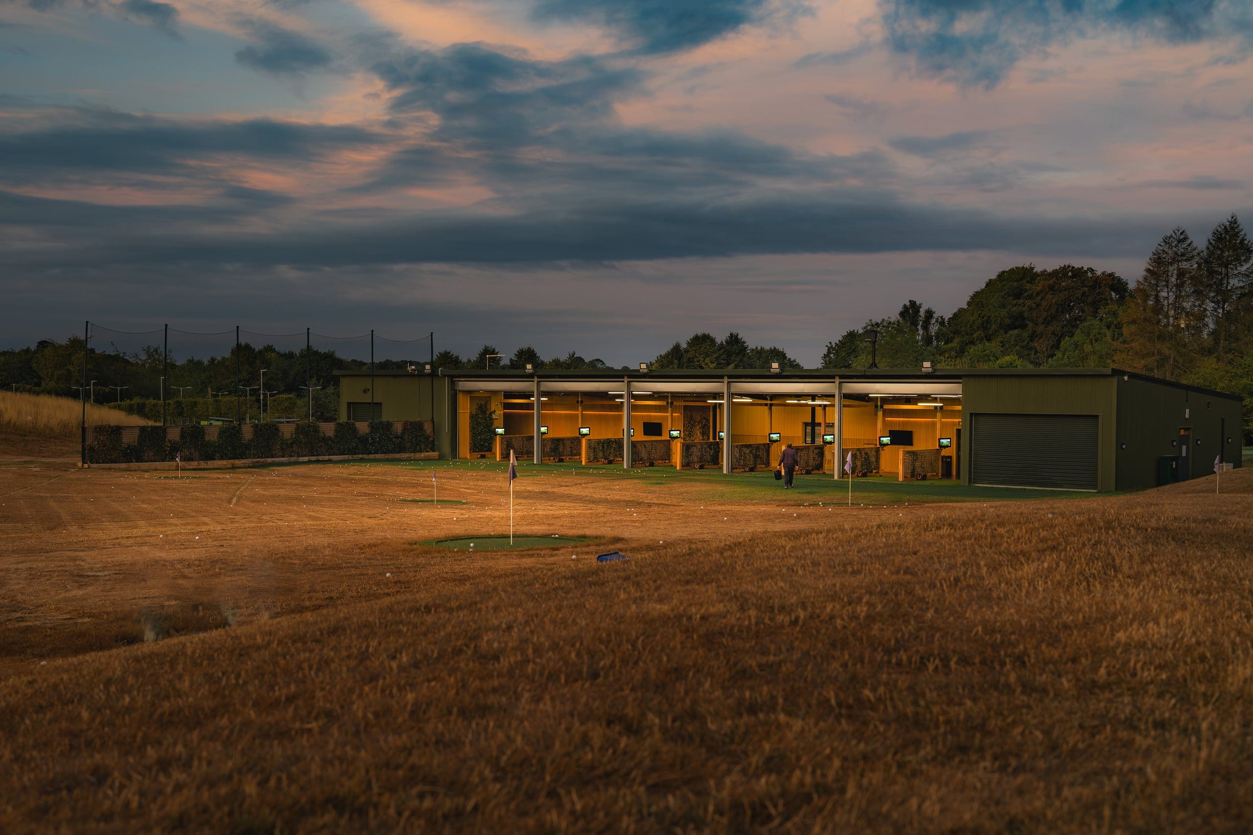 The floodlit driving range bays at dusk, the practice green in the foreground