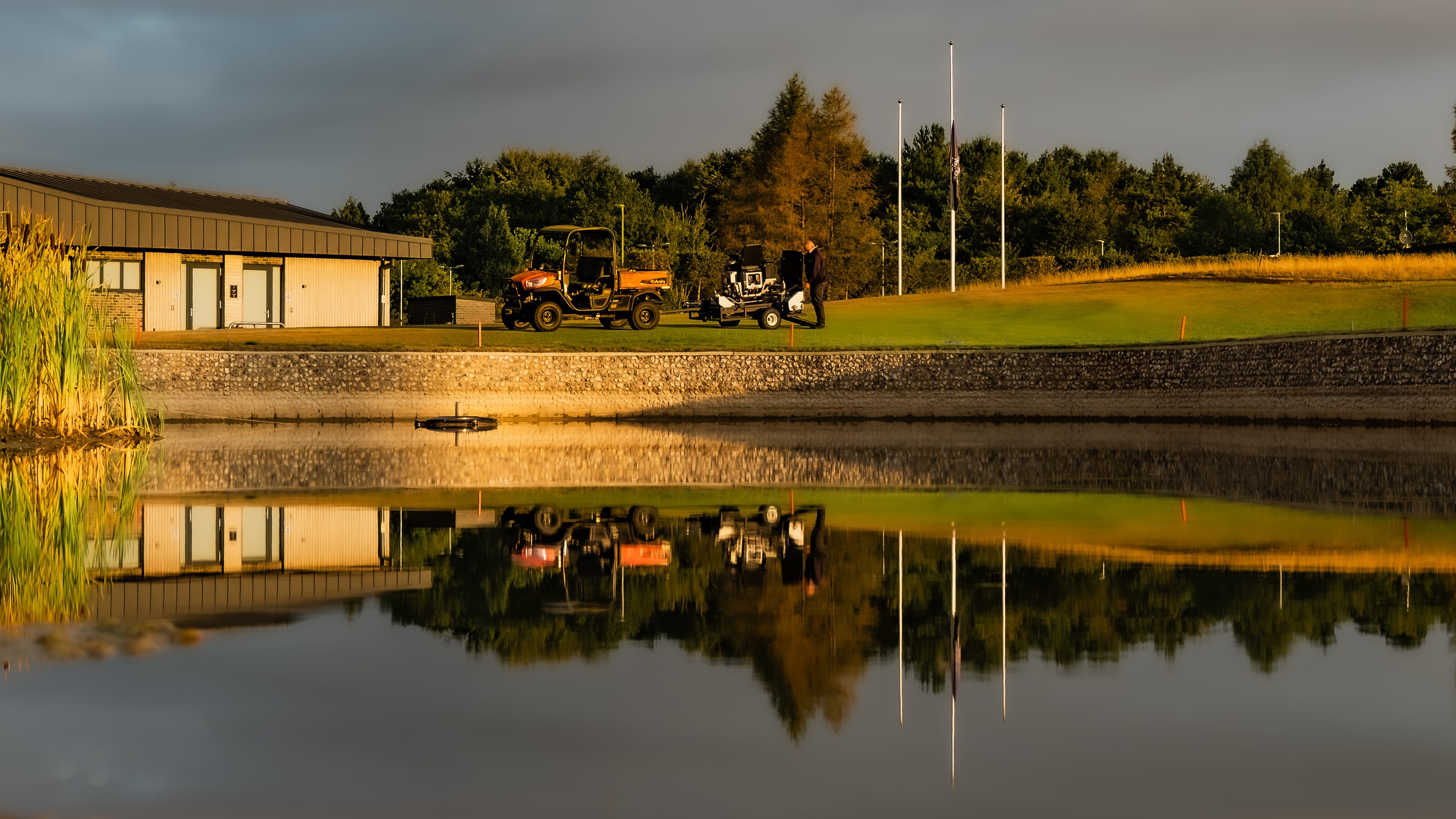 Greenkeepers loading equipment onto a utility vehicle beside the practice pond, the clubhouse reflected in the water