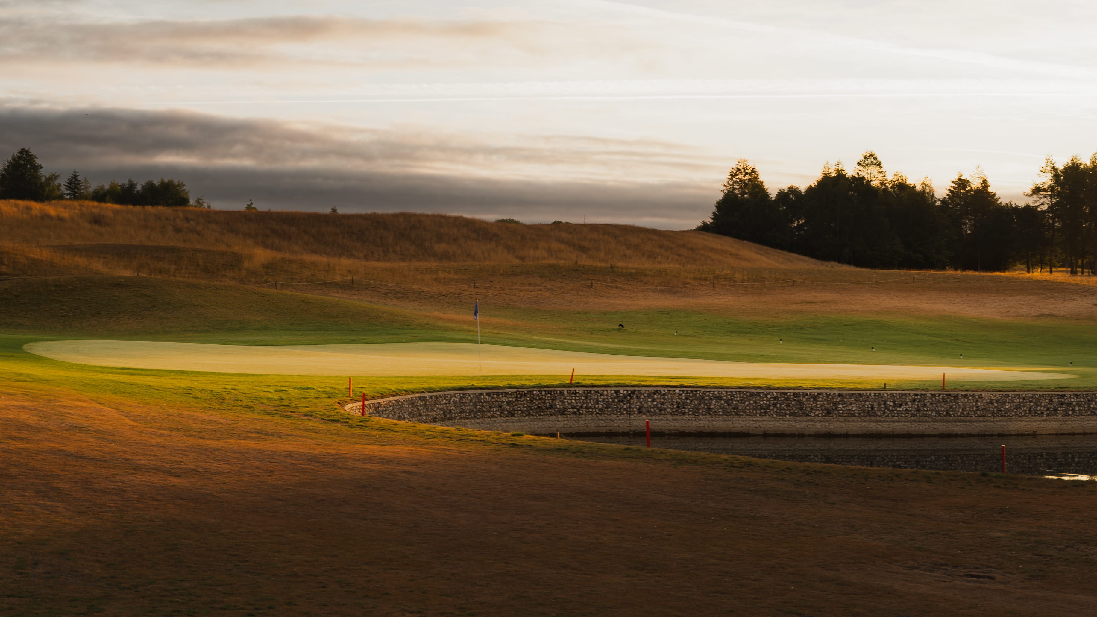 A green and bunker lit by low evening light, mist over the fairway beyond