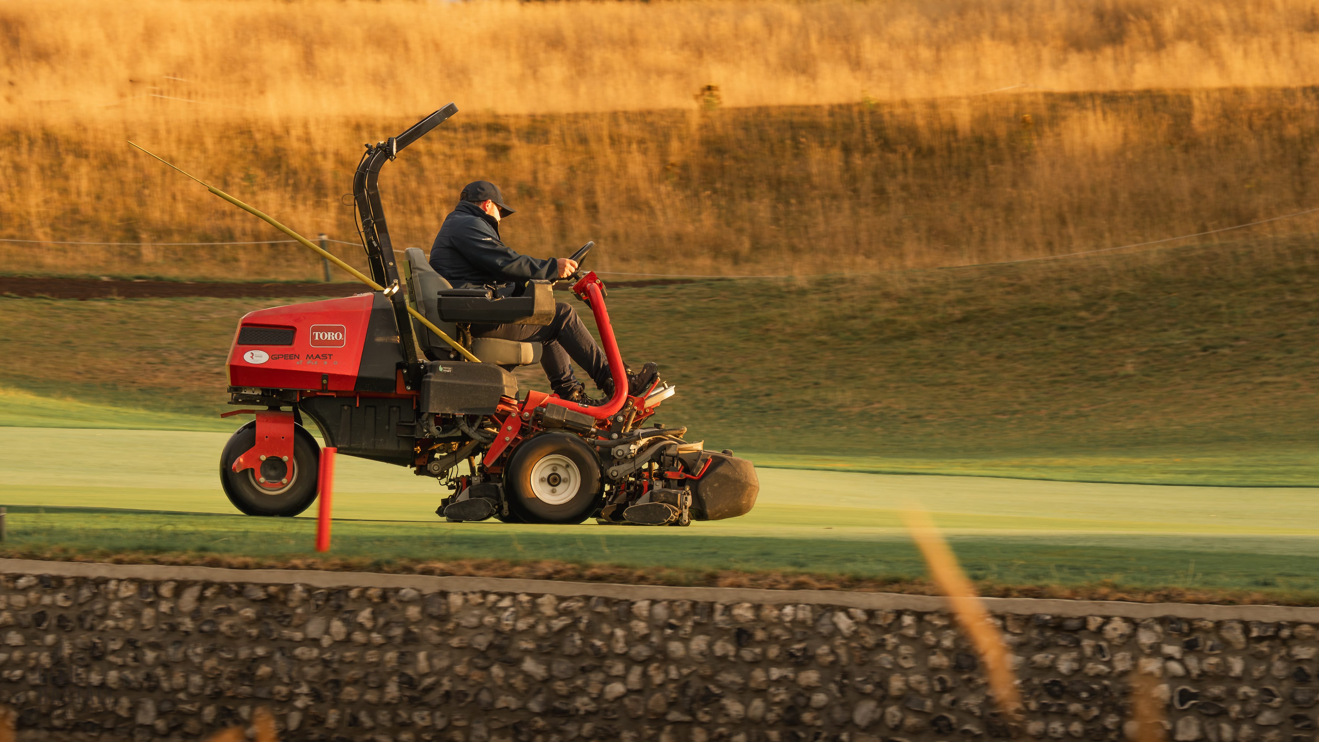 A greenkeeper on a triple mower cutting a green in low golden light