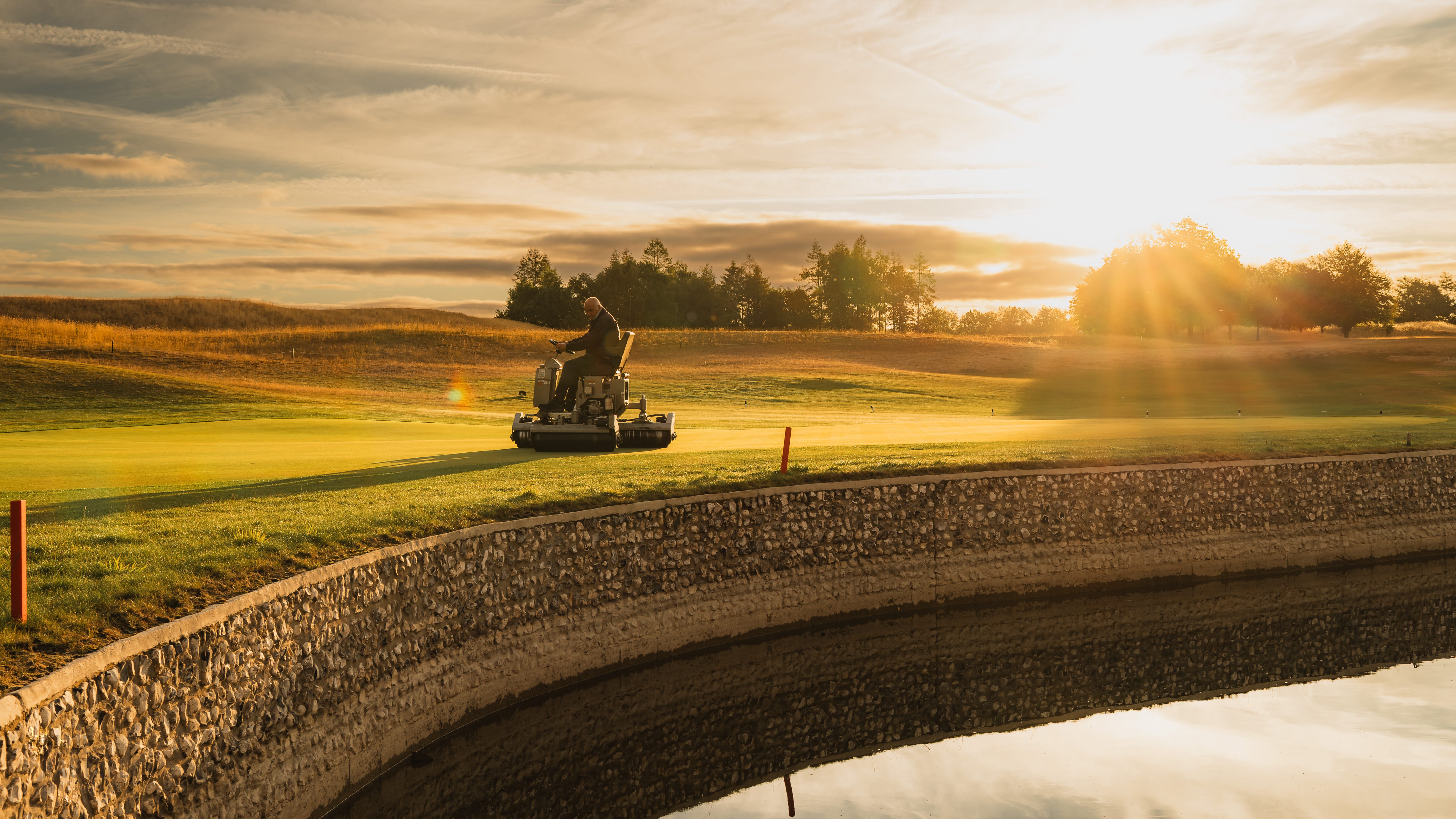 A mower working beside a stone-lined bank at golden hour