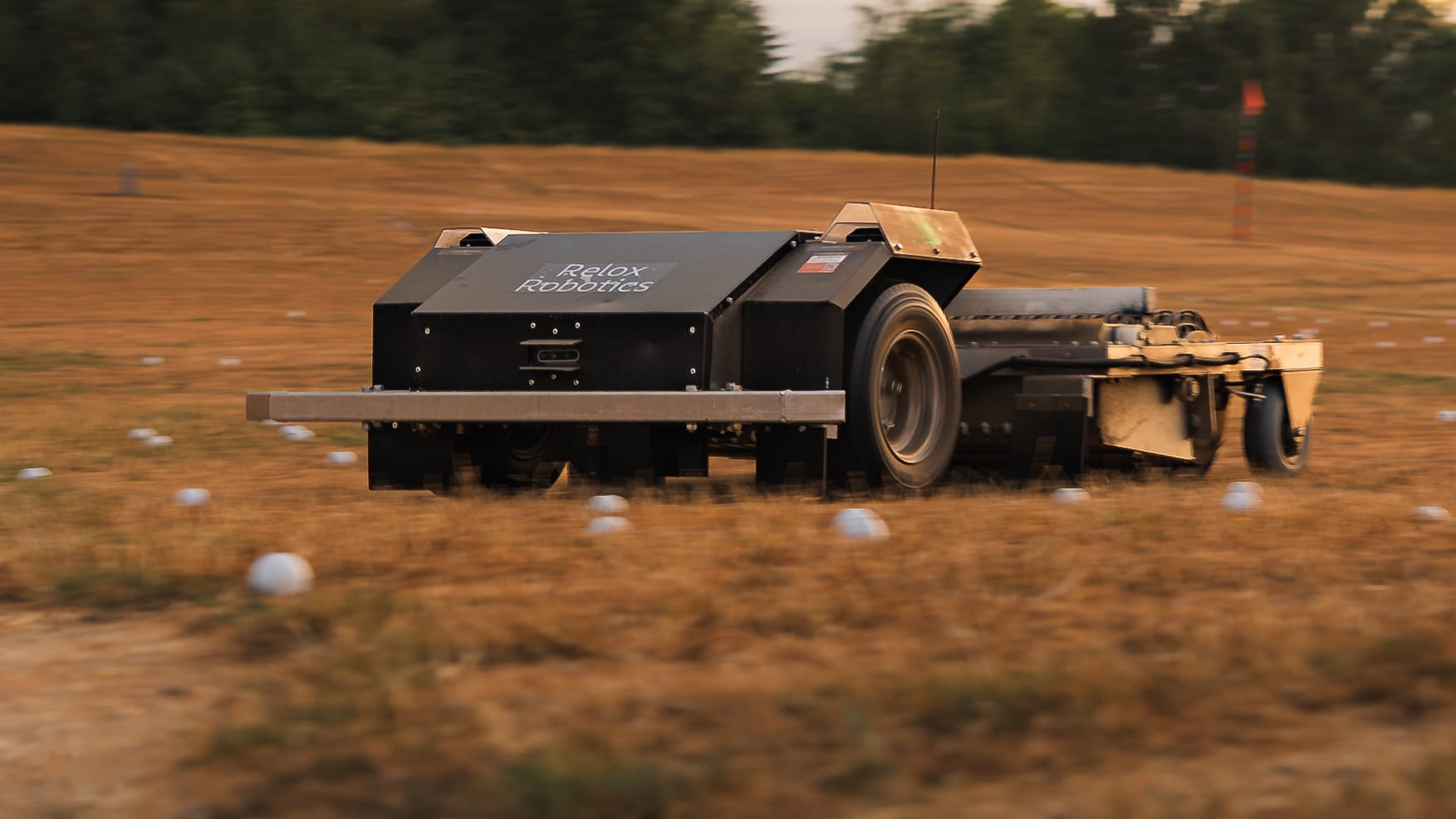 An autonomous Relox Robotics ball collector crossing the driving range, golf balls scattered across the grass