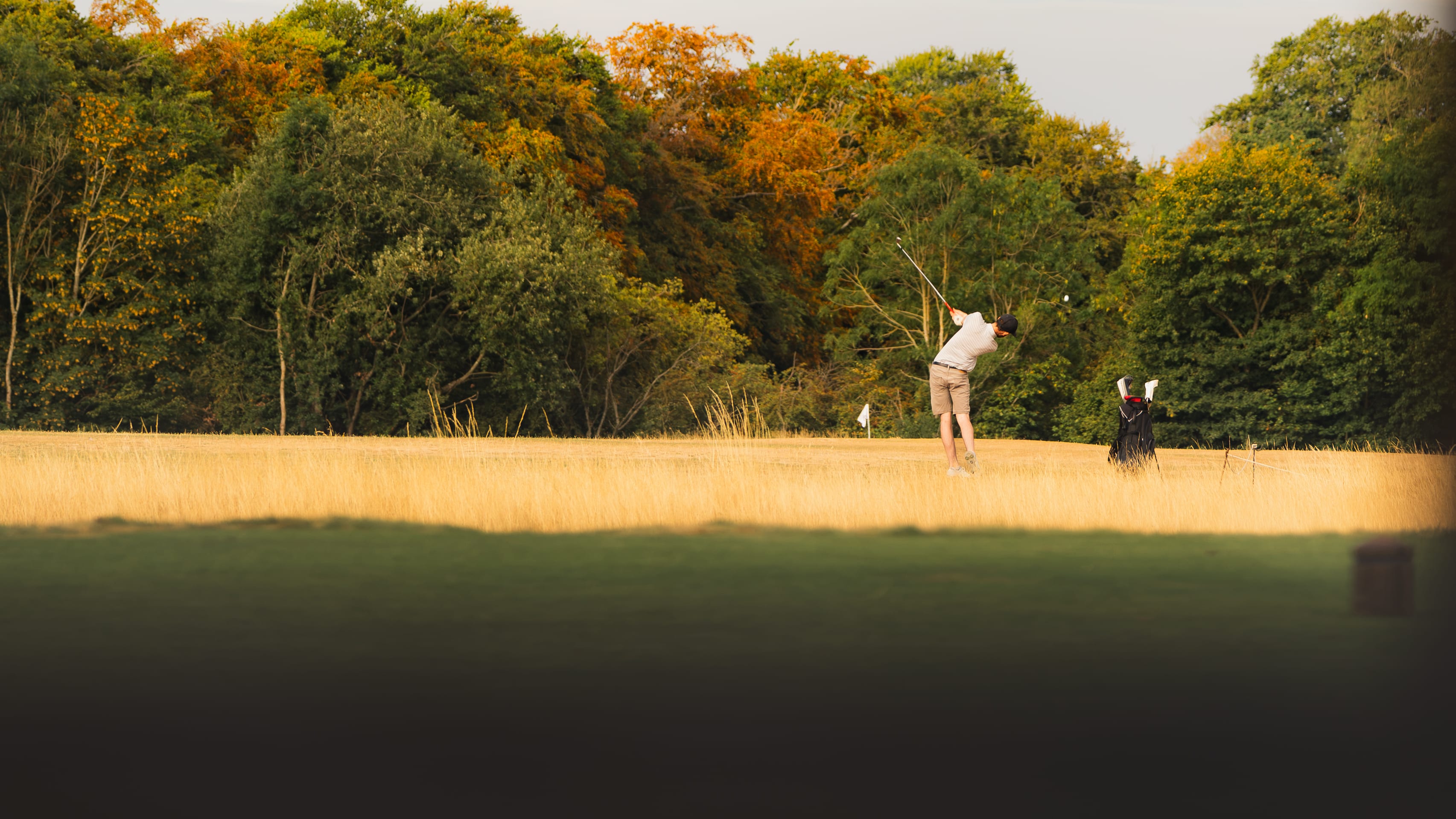 A golfer mid-swing on the fairway, autumn colour in the trees behind