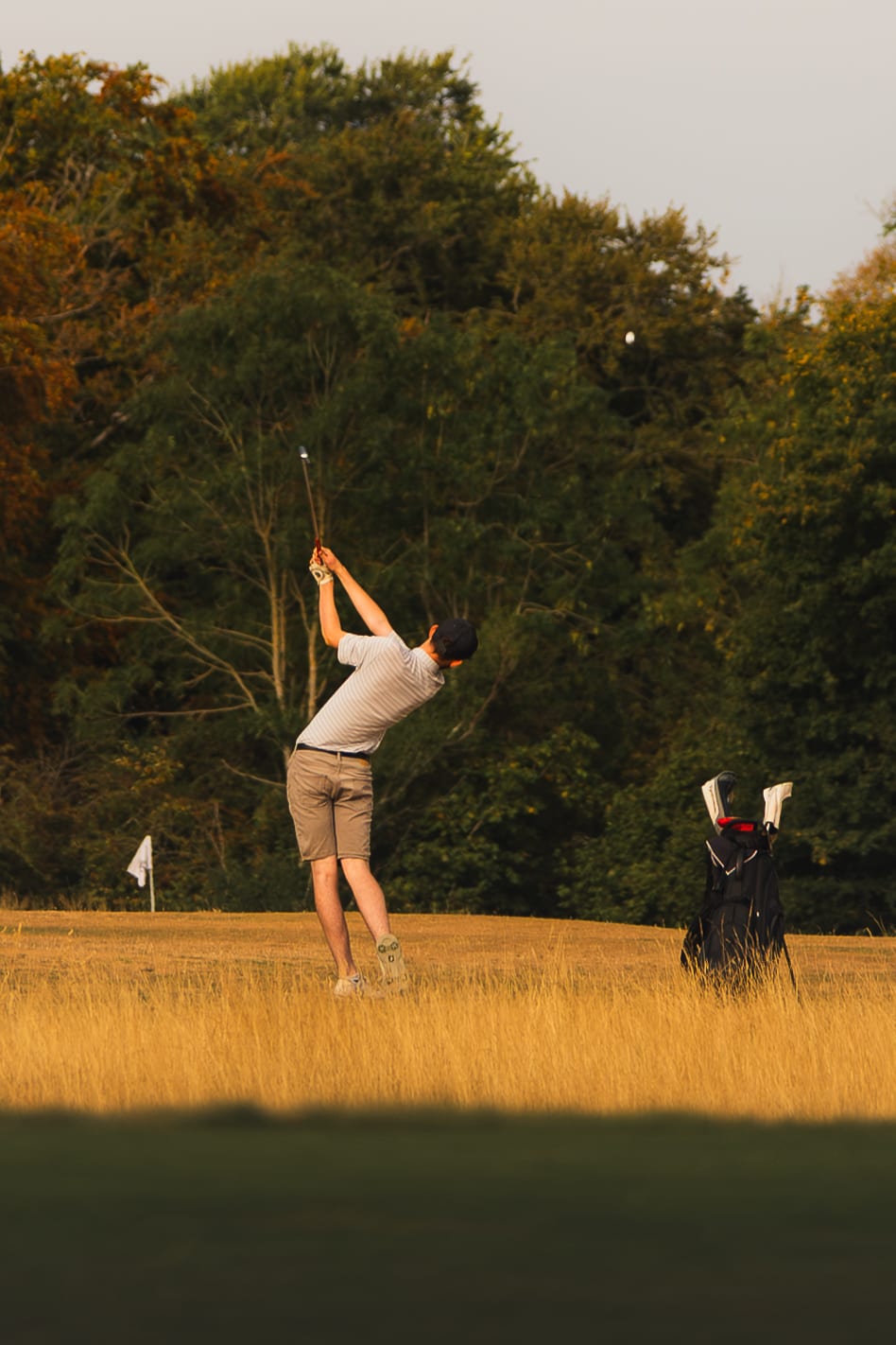 A golfer completing his swing at sunset, clubs standing beside him