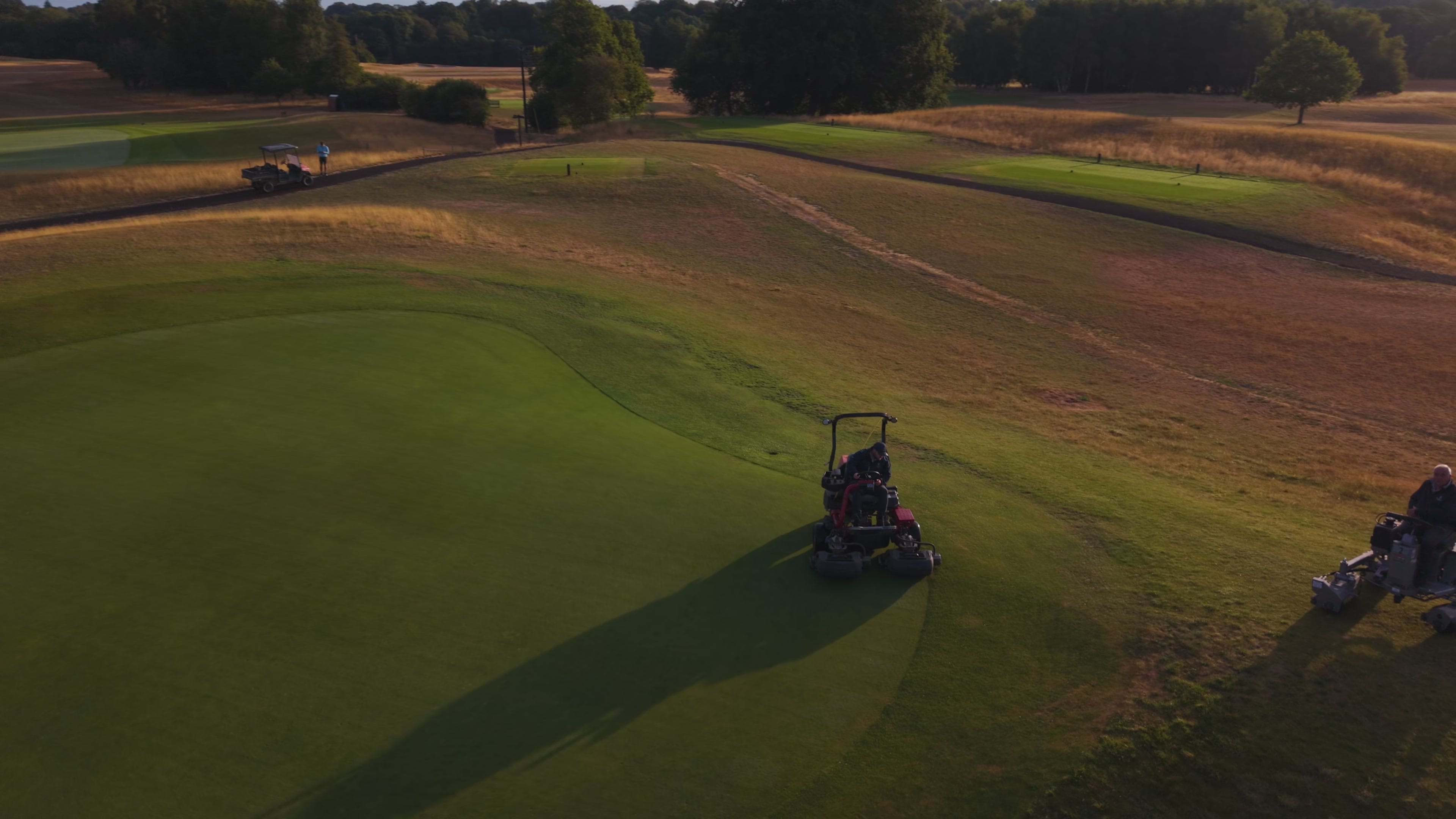 A triple mower cutting a green at golden hour, its shadow thrown across the turf