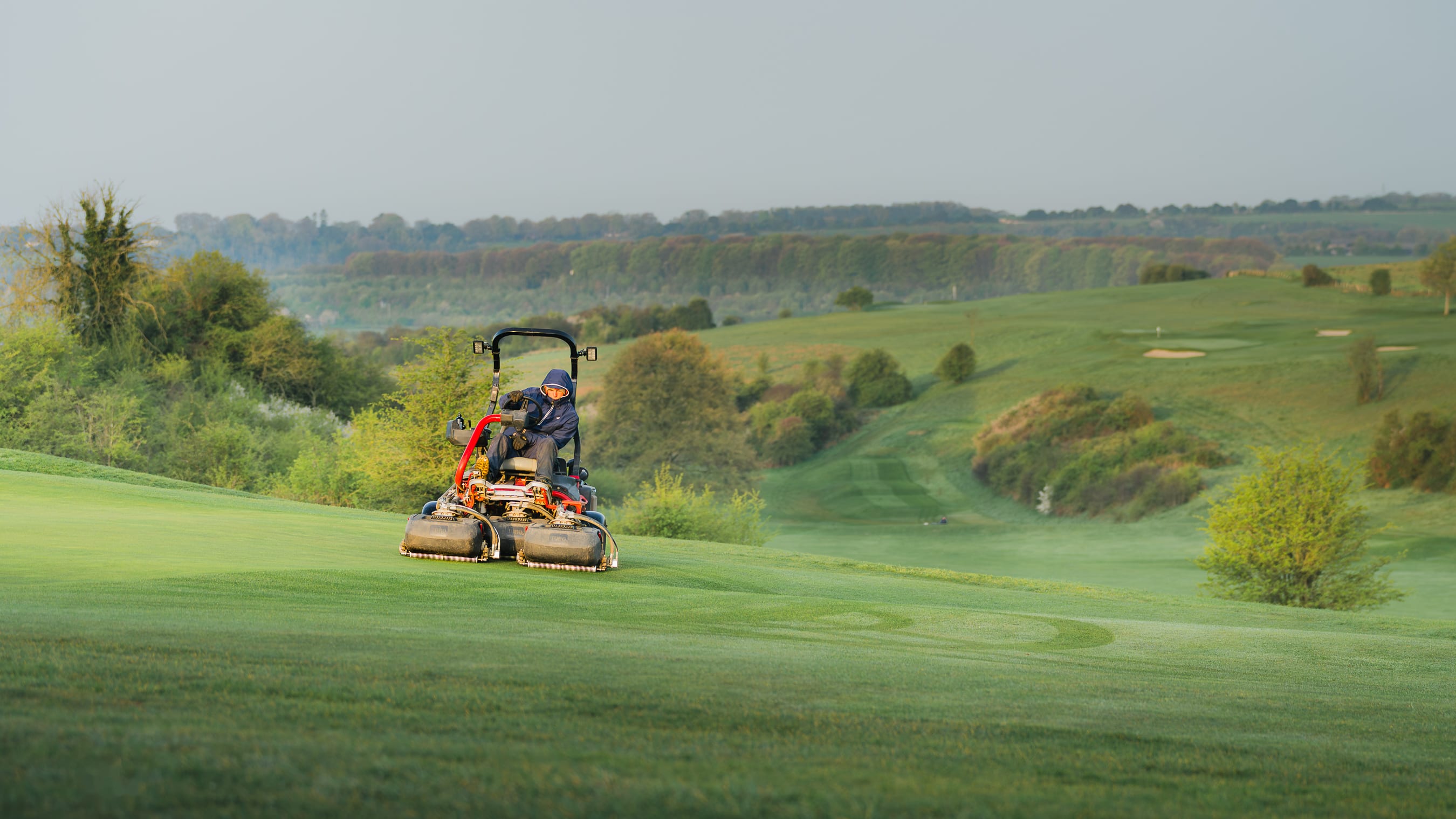 A greenkeeper on a triple mower cutting a fairway in early morning light, rolling countryside beyond