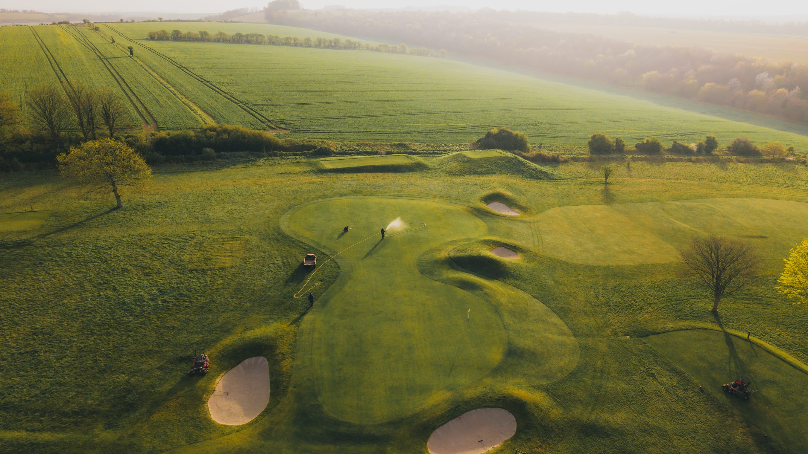 Aerial view through morning mist of a green being watered by two greenkeepers, bunkers either side