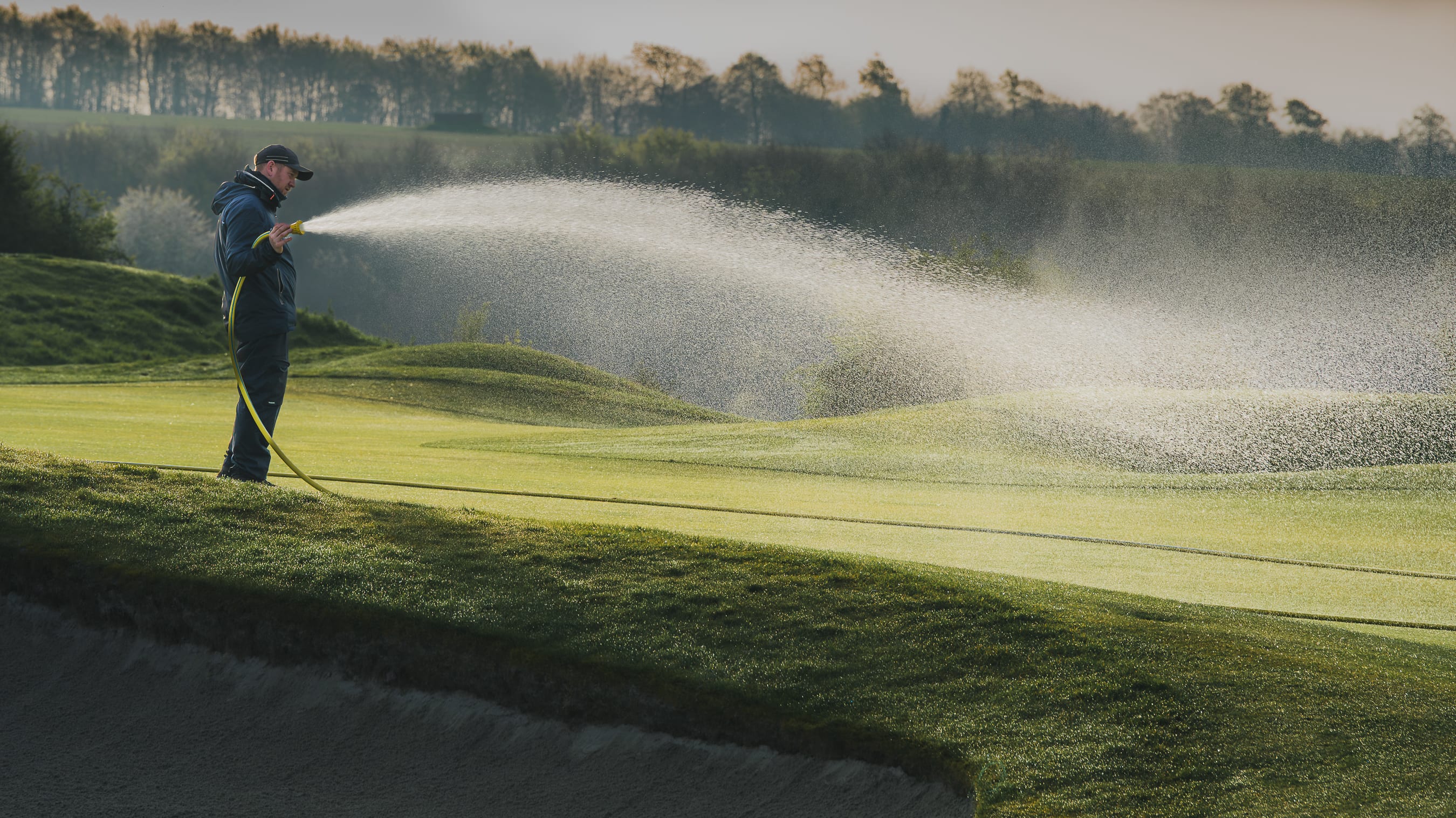 Two greenkeepers working a green at dawn, one hand-watering and one on a mower, rolling hills behind