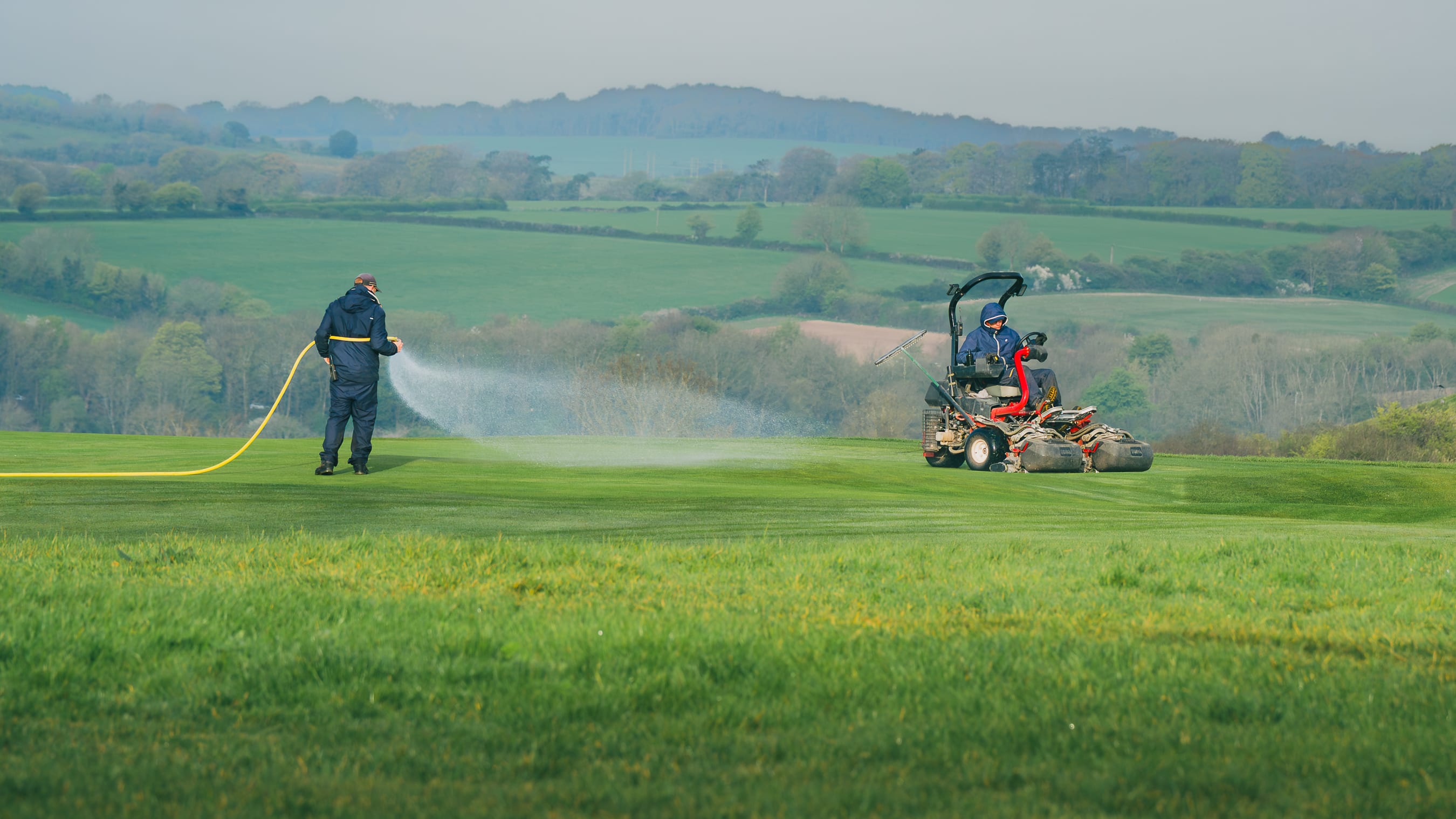 A greenkeeper on a triple mower silhouetted against the Hampshire countryside