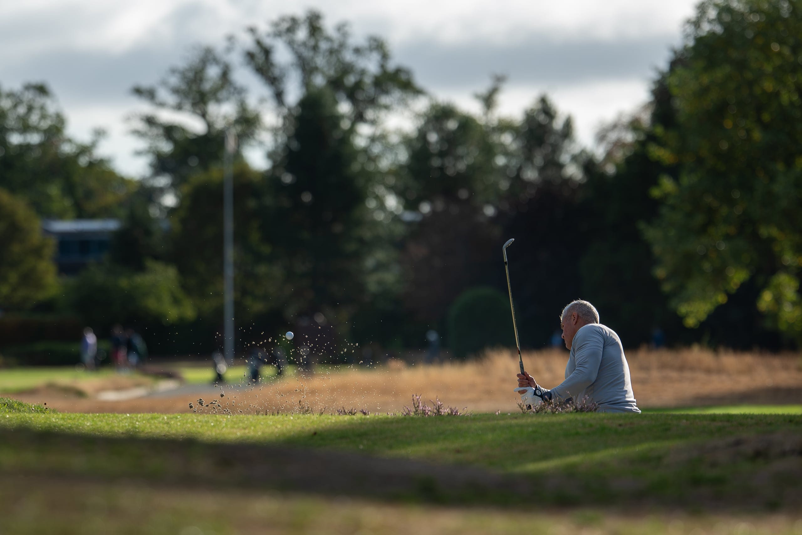 A golfer playing out of a bunker, spectators watching from beyond the green