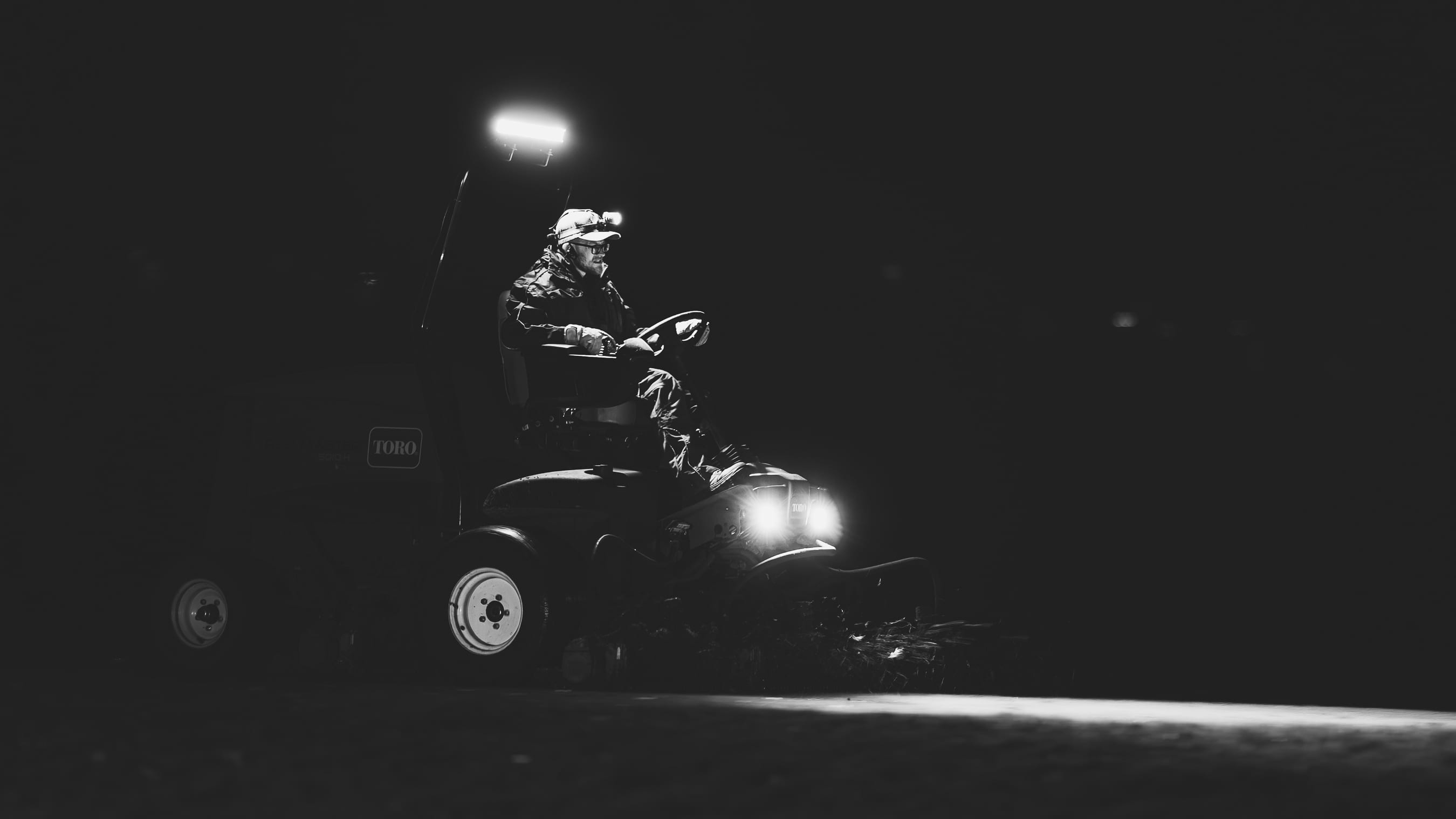 A greenkeeper on a Toro mower working a green in the dark, the machine lit by its own work lights