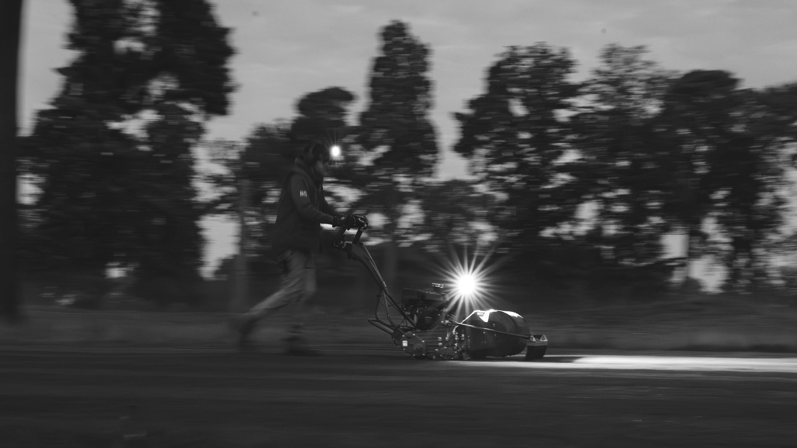 A greenkeeper walking a pedestrian mower across a green at first light, in black and white