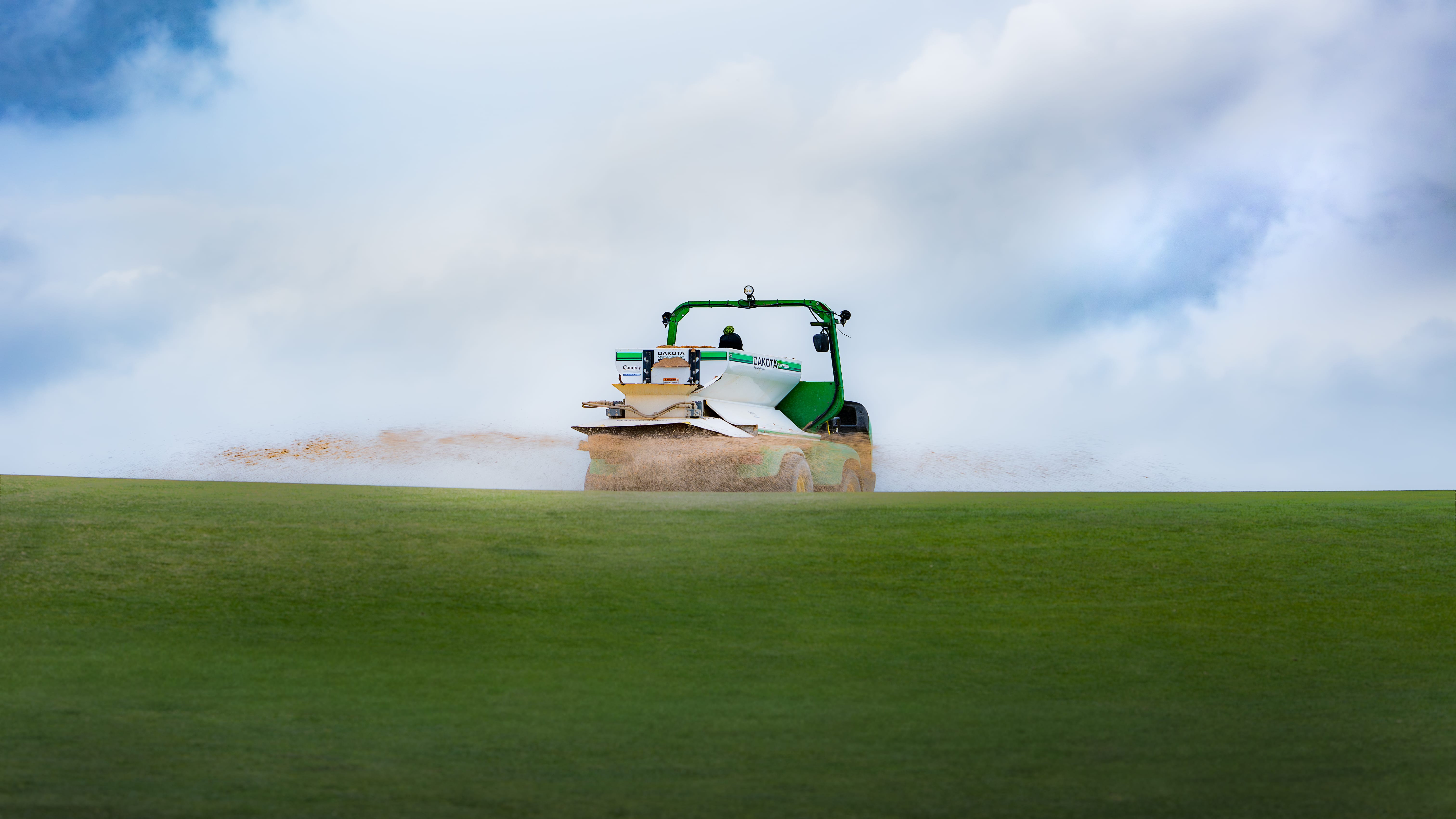 A sand spreader topdressing a green, the material caught in the air against a bank of cloud