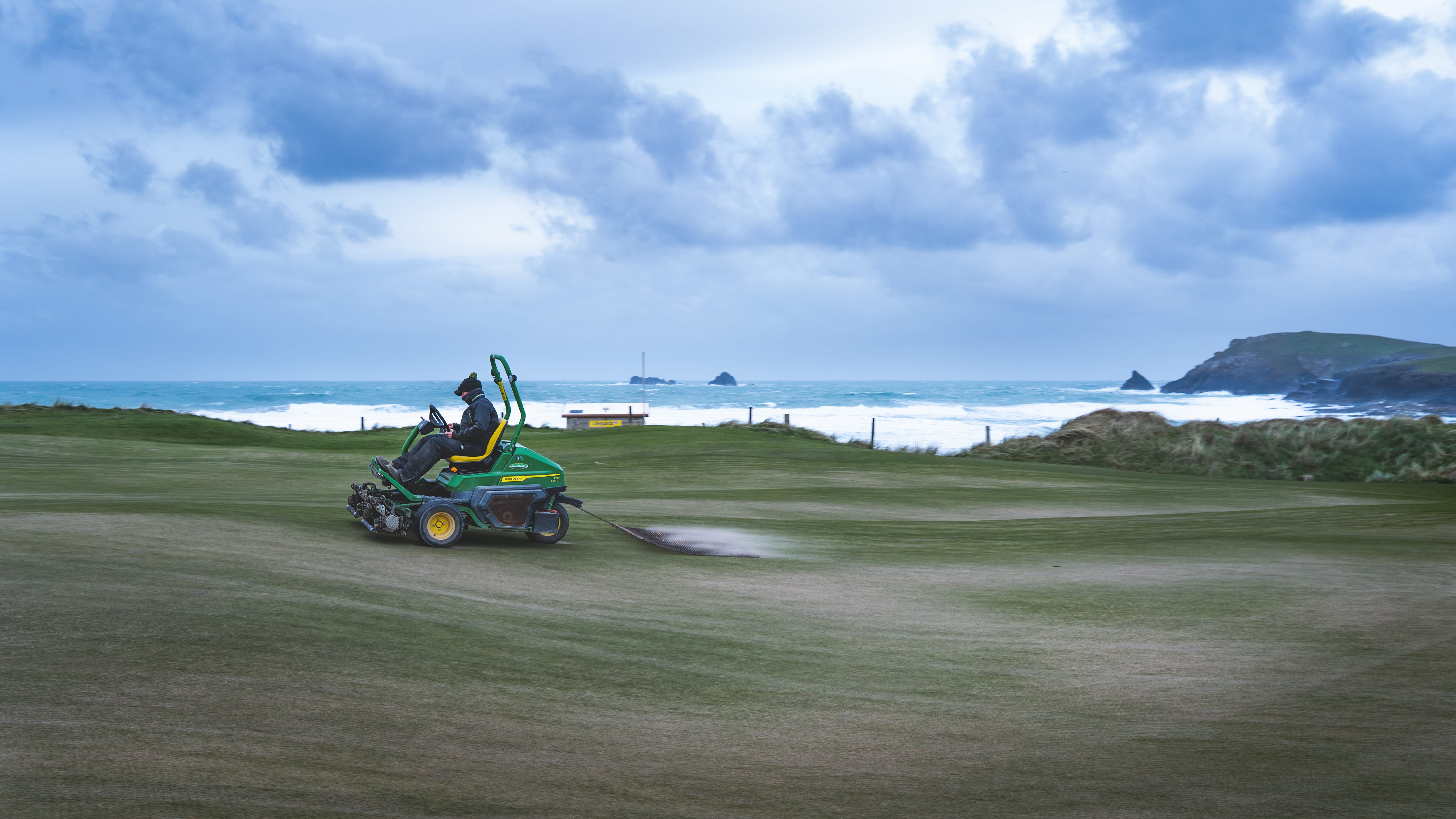 A greenkeeper dragging sand into a green, the sea and cliffs beyond