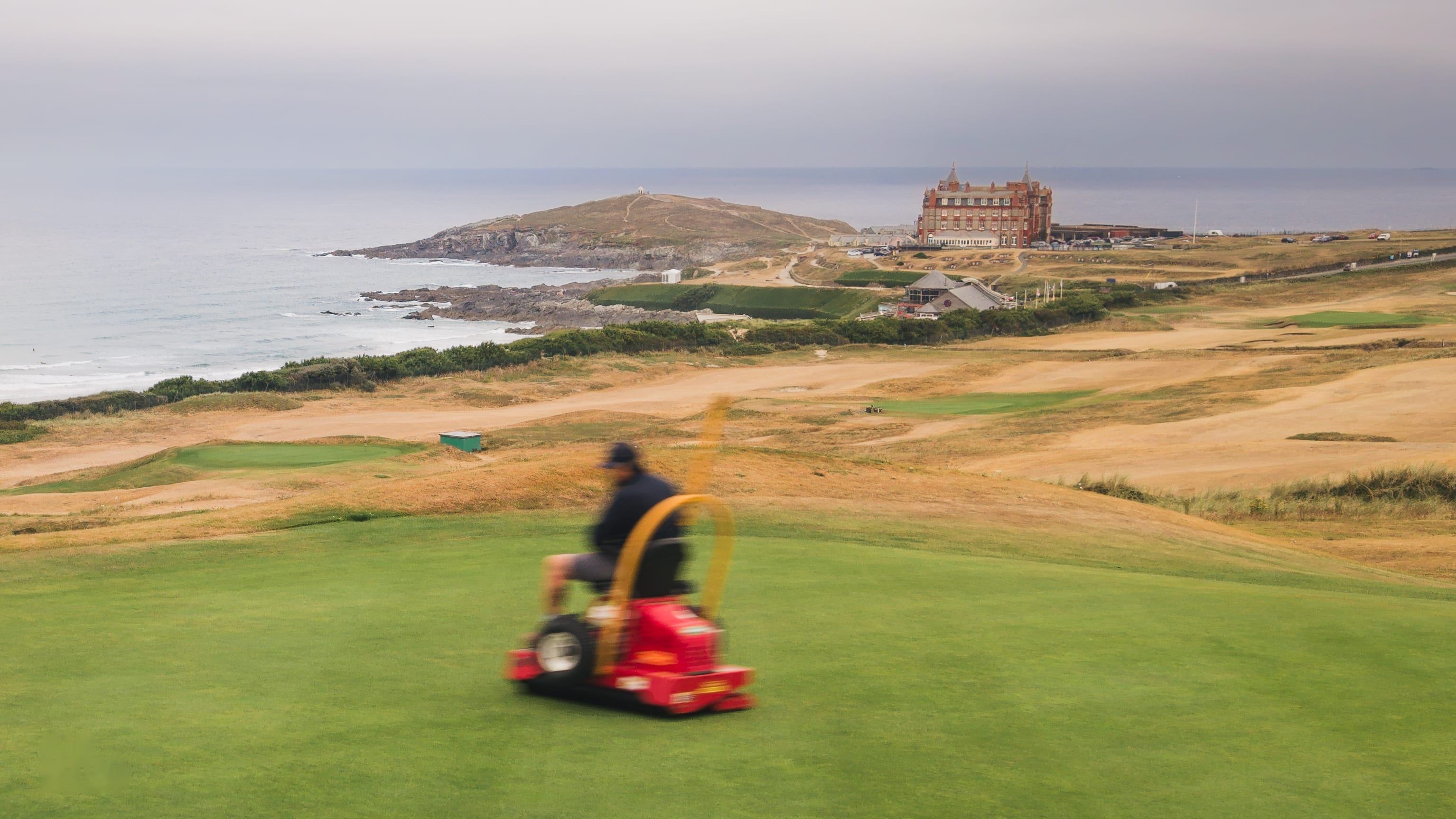 A red Tru-Turf roller working a green above the sea at Newquay, the headland hotel beyond